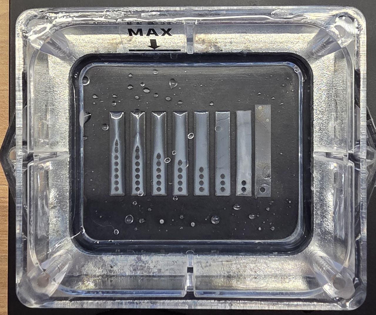 A real exposure test strip cured in the vat: eight bars with dot markers, holes crisp on the long-exposure side and sealing over toward the short side